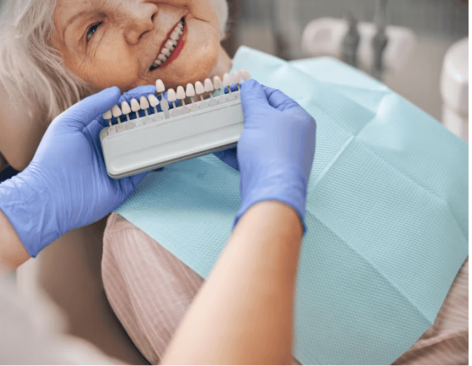 A dentist wearing gloves holds a set of dental shade guides next to an older woman’s smile as she sits in a dental chair, wearing a blue protective bib.