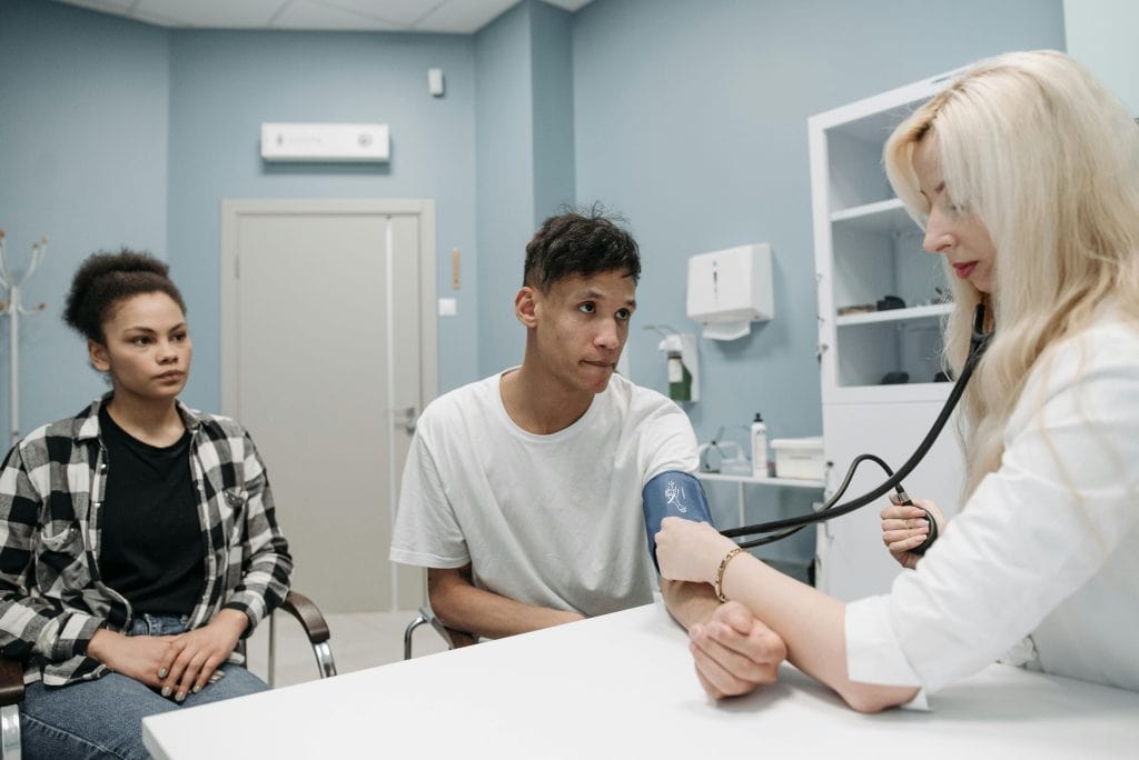 Empathy in healthcare: doctor measures blood pressure of young male patient while a young woman observes in waiting room.